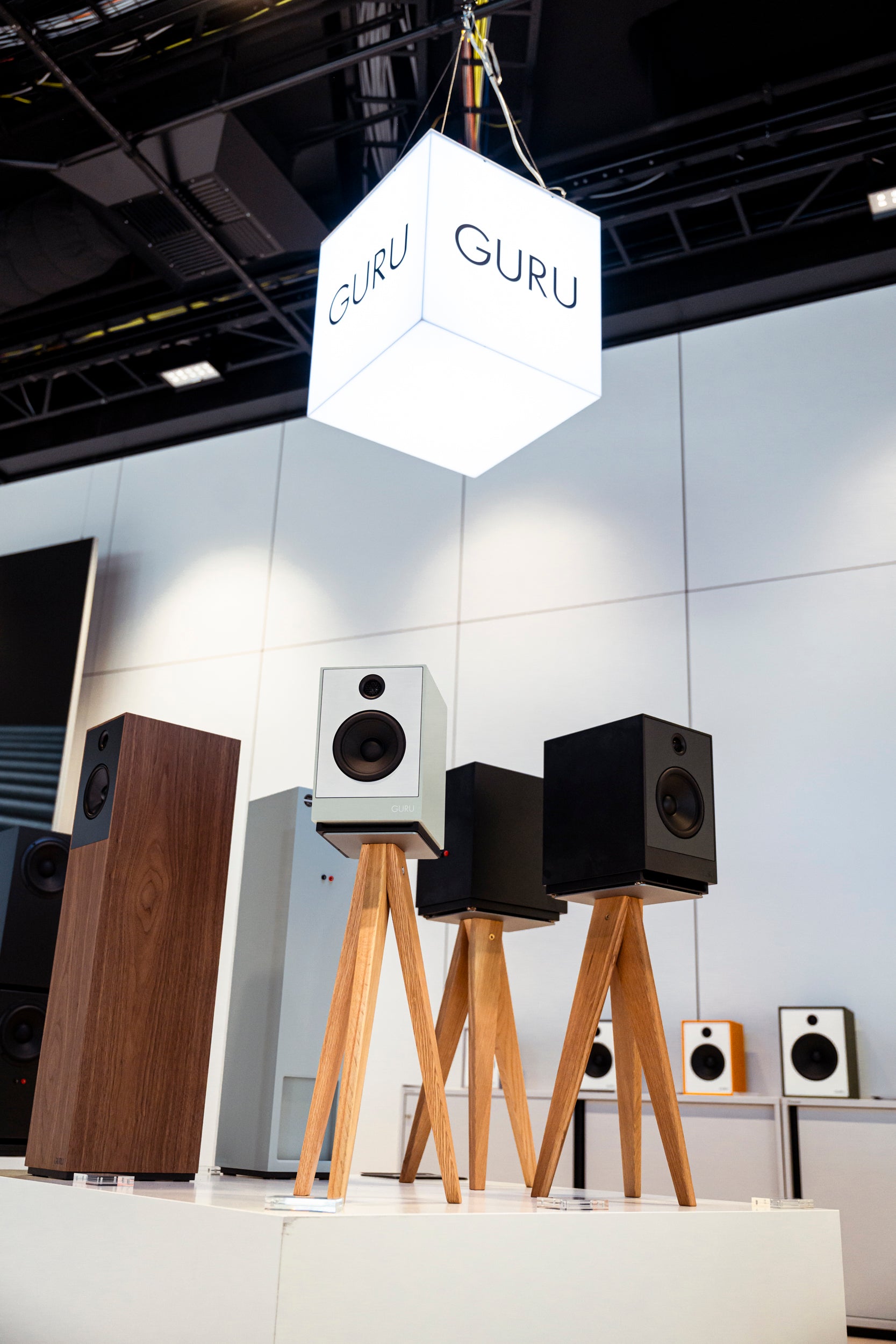 A display of GURU brand speakers at an exhibition, featuring two white and two black speakers on wooden tripod stands under a hanging white cube sign with the GURU logo.